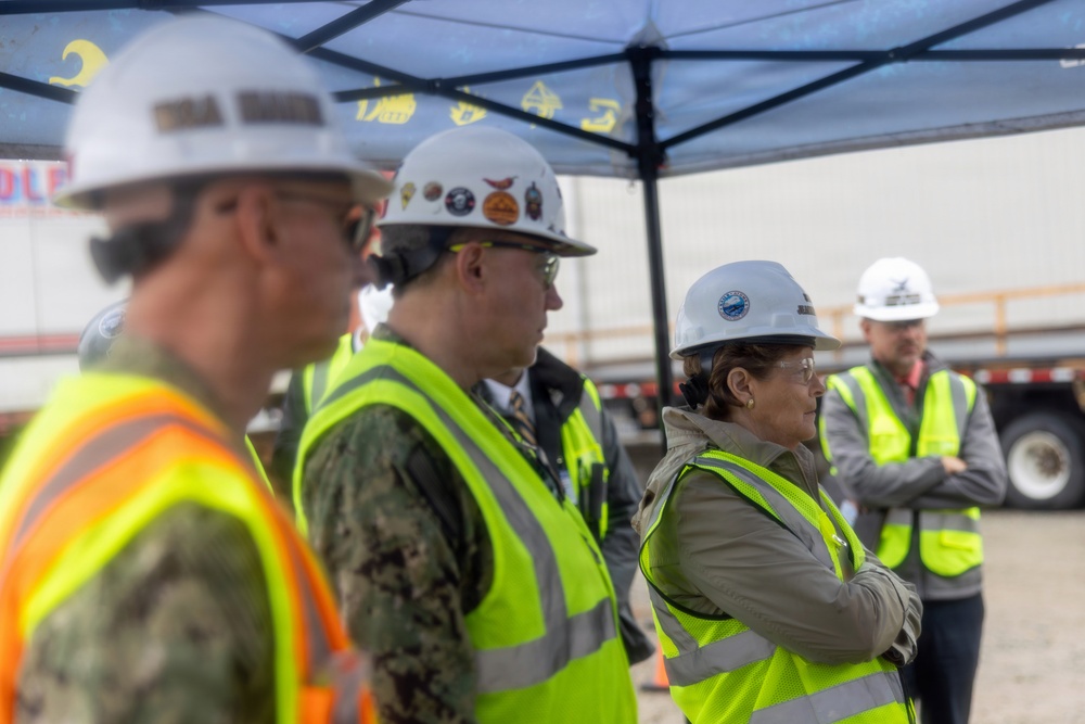 A group of individuals wearing hard hats and high-visibility safety vests are gathered at what appears to be a construction or industrial site. At least one person is wearing a military uniform (Navy digital camouflage), suggesting a joint civilian-military site visit or inspection. The group appears to be observing or listening to something outside the frame.