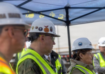 A group of individuals wearing hard hats and high-visibility safety vests are gathered at what appears to be a construction or industrial site. At least one person is wearing a military uniform (Navy digital camouflage), suggesting a joint civilian-military site visit or inspection. The group appears to be observing or listening to something outside the frame.