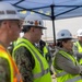 A group of individuals wearing hard hats and high-visibility safety vests are gathered at what appears to be a construction or industrial site. At least one person is wearing a military uniform (Navy digital camouflage), suggesting a joint civilian-military site visit or inspection. The group appears to be observing or listening to something outside the frame.