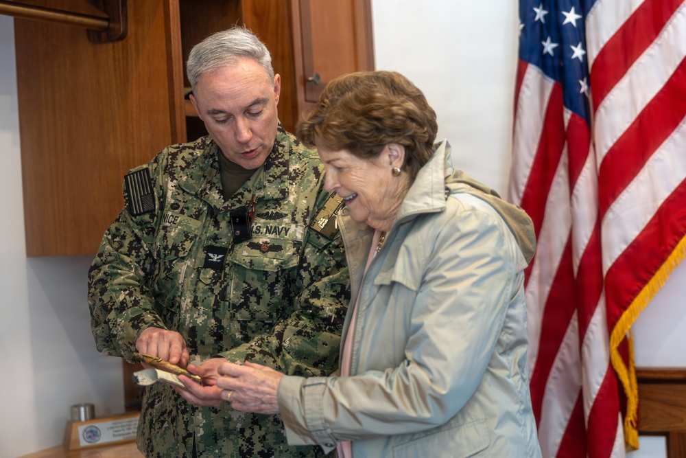 A U.S. Navy officer in digital camouflage uniform is showing or presenting a small item to a civilian woman in an indoor official setting. Both individuals are looking down at the object being exchanged or examined. An American flag with gold fringe is prominently displayed in the background.