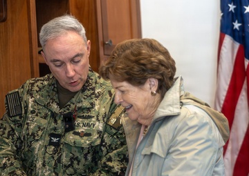 A U.S. Navy officer in digital camouflage uniform is showing or presenting a small item to a civilian woman in an indoor official setting. Both individuals are looking down at the object being exchanged or examined. An American flag with gold fringe is prominently displayed in the background.