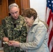A U.S. Navy officer in digital camouflage uniform is showing or presenting a small item to a civilian woman in an indoor official setting. Both individuals are looking down at the object being exchanged or examined. An American flag with gold fringe is prominently displayed in the background.
