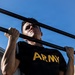 A male individual wearing a black U.S. Army physical training t-shirt is performing pull-ups on an outdoor bar structure. The photo is taken from a low angle looking upward, with a clear blue sky in the background. The subject appears to be mid-exercise, with visible perspiration and muscular exertion.