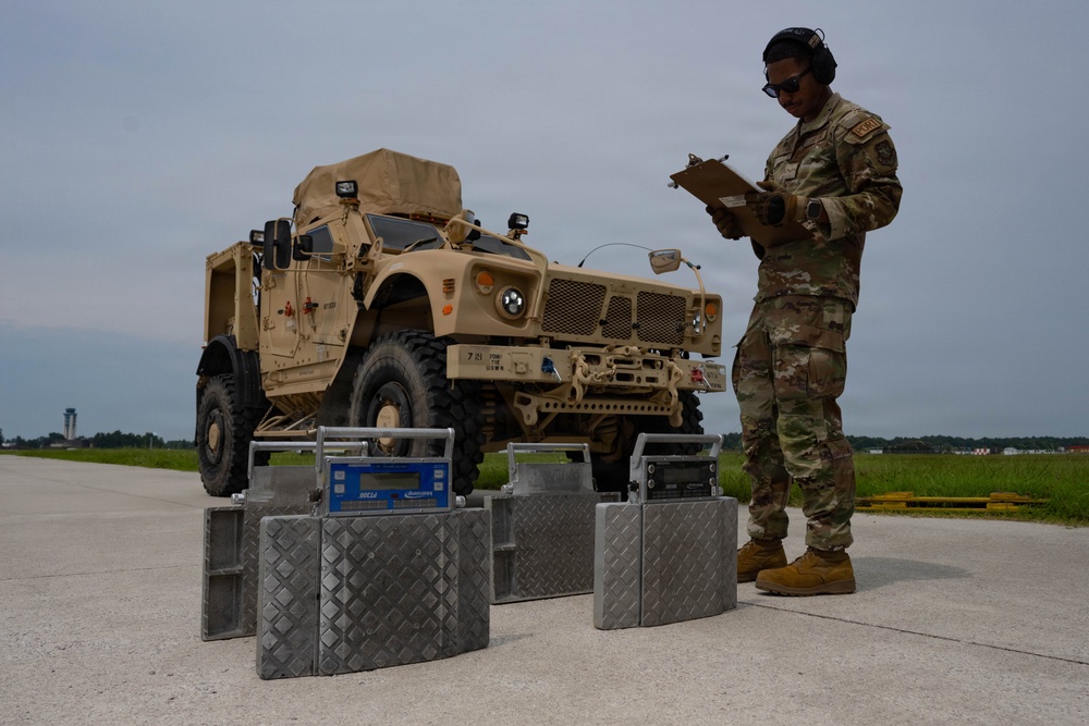 A military service member in OCP (Operational Camouflage Pattern) uniform is recording data on a clipboard while standing next to portable axle/wheel weighing scales positioned in front of a tan/desert-colored MRAP or similar armored military vehicle. The activity appears to be a vehicle weight and balance inspection, likely conducted on an airfield tarmac in preparation for air transport. The setting suggests a U.S. military installation with an air traffic control tower visible in the background.