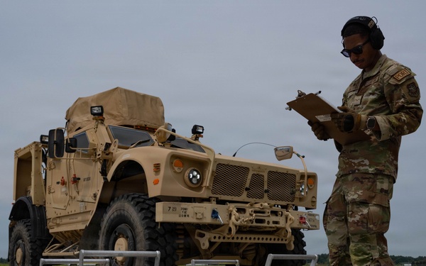 A military service member in OCP (Operational Camouflage Pattern) uniform is recording data on a clipboard while standing next to portable axle/wheel weighing scales positioned in front of a tan/desert-colored MRAP or similar armored military vehicle. The activity appears to be a vehicle weight and balance inspection, likely conducted on an airfield tarmac in preparation for air transport. The setting suggests a U.S. military installation with an air traffic control tower visible in the background.