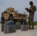 A military service member in OCP (Operational Camouflage Pattern) uniform is recording data on a clipboard while standing next to portable axle/wheel weighing scales positioned in front of a tan/desert-colored MRAP or similar armored military vehicle. The activity appears to be a vehicle weight and balance inspection, likely conducted on an airfield tarmac in preparation for air transport. The setting suggests a U.S. military installation with an air traffic control tower visible in the background.