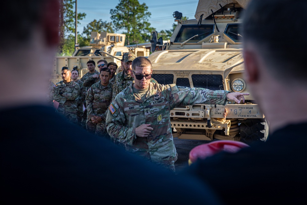 A U.S. Army soldier, identified by name tape as 'RIOS,' is conducting a briefing or instruction session for a group of fellow soldiers in front of military vehicles. The soldier is gesturing with his right arm, appearing to point toward or reference the vehicle behind him. The setting appears to be an outdoor motor pool or staging area with multiple tan/desert-colored military tactical vehicles.