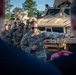 A U.S. Army soldier, identified by name tape as 'RIOS,' is conducting a briefing or instruction session for a group of fellow soldiers in front of military vehicles. The soldier is gesturing with his right arm, appearing to point toward or reference the vehicle behind him. The setting appears to be an outdoor motor pool or staging area with multiple tan/desert-colored military tactical vehicles.