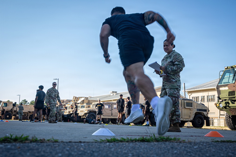 A military physical fitness test is being conducted in an outdoor area of what appears to be a U.S. Army installation. A soldier in PT (physical training) attire is sprinting past a female soldier in Army Combat Uniform (ACU/OCP) who is observing and recording results on a clipboard. Multiple military vehicles including HMMWVs (Humvees) and larger tactical trucks are parked in the background.
