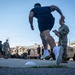 A military physical fitness test is being conducted in an outdoor area of what appears to be a U.S. Army installation. A soldier in PT (physical training) attire is sprinting past a female soldier in Army Combat Uniform (ACU/OCP) who is observing and recording results on a clipboard. Multiple military vehicles including HMMWVs (Humvees) and larger tactical trucks are parked in the background.