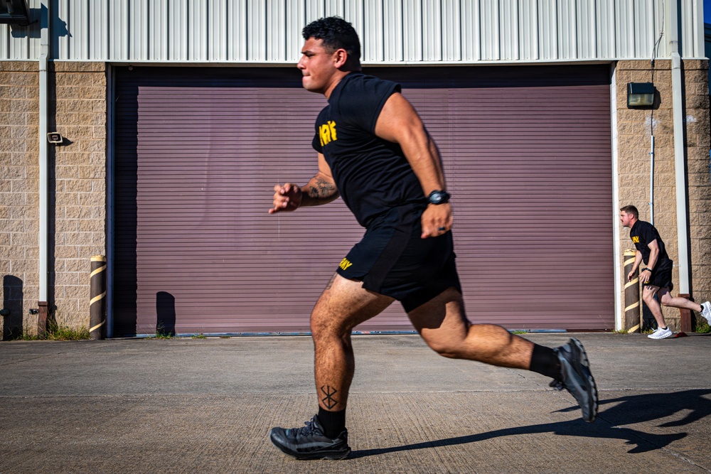 A muscular man in black military-style PT (physical training) gear is captured mid-sprint on a paved surface in front of an industrial building with a large roll-up door. A second person in identical black and yellow PT uniform is visible in the background also running. The clothing bears yellow text consistent with U.S. Army physical training uniforms.