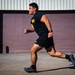 A muscular man in black military-style PT (physical training) gear is captured mid-sprint on a paved surface in front of an industrial building with a large roll-up door. A second person in identical black and yellow PT uniform is visible in the background also running. The clothing bears yellow text consistent with U.S. Army physical training uniforms.