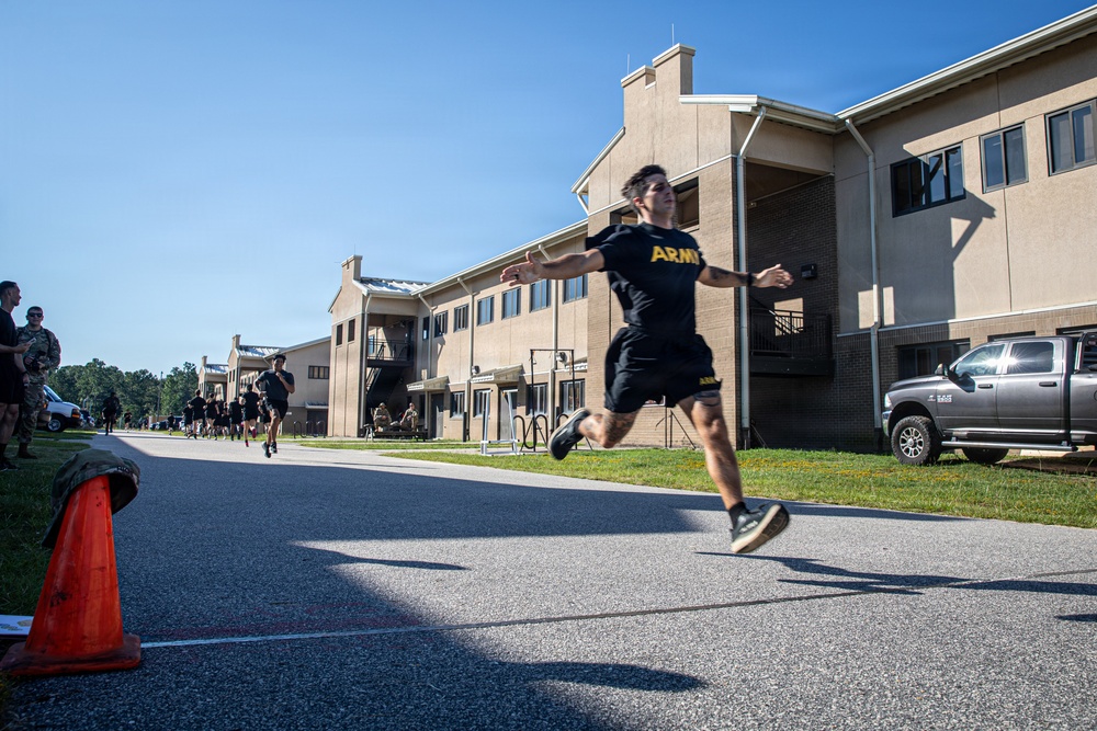 A soldier wearing U.S. Army physical training (PT) uniform is captured mid-leap on a paved road alongside military barracks-style buildings. Additional soldiers in matching black Army PT uniforms are running in the background, suggesting a group physical fitness event or run. The image is taken at a military installation during daytime under clear blue skies.