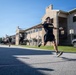 A soldier wearing U.S. Army physical training (PT) uniform is captured mid-leap on a paved road alongside military barracks-style buildings. Additional soldiers in matching black Army PT uniforms are running in the background, suggesting a group physical fitness event or run. The image is taken at a military installation during daytime under clear blue skies.