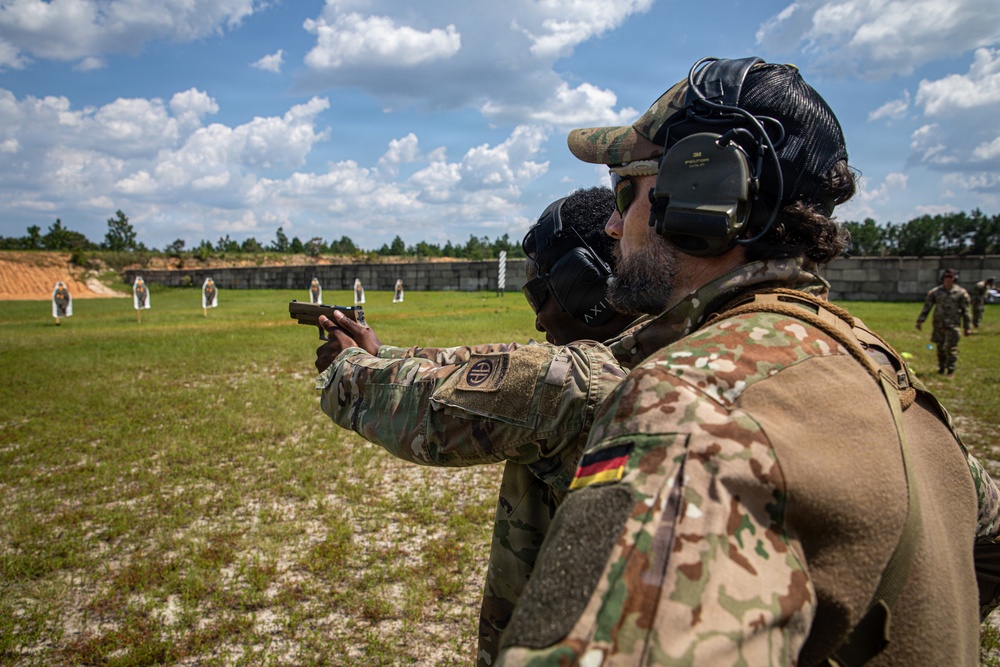 Two military personnel are at an outdoor shooting range, with one soldier aiming a pistol downrange at paper targets while another observes closely from behind. The soldier in the foreground wears a German flag patch on their uniform, while the shooter displays an 82nd Airborne Division patch. Both are wearing hearing protection and are in full military camouflage uniforms.