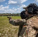 Two military personnel are at an outdoor shooting range, with one soldier aiming a pistol downrange at paper targets while another observes closely from behind. The soldier in the foreground wears a German flag patch on their uniform, while the shooter displays an 82nd Airborne Division patch. Both are wearing hearing protection and are in full military camouflage uniforms.