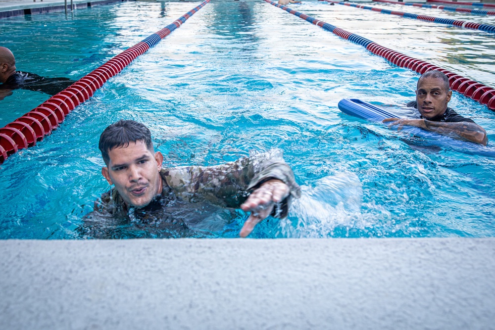 Three individuals are in a competitive swimming pool with red and blue lane dividers. The central figure is wearing camouflage military uniform while swimming, suggesting military water survival or combat swimmer training. A second person on the right uses a blue kickboard, while a third person is partially visible on the left.