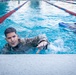 Three individuals are in a competitive swimming pool with red and blue lane dividers. The central figure is wearing camouflage military uniform while swimming, suggesting military water survival or combat swimmer training. A second person on the right uses a blue kickboard, while a third person is partially visible on the left.