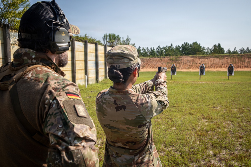 Two military personnel are at an outdoor shooting range. A soldier identified by name tape as 'SUAZO' is firing a pistol at humanoid silhouette targets downrange, while a German military observer wearing a German flag patch and tactical hearing protection stands nearby. The setting appears to be a formal military training range with a berm backstop and wooden barrier wall.