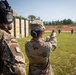 Two military personnel are at an outdoor shooting range. A soldier identified by name tape as 'SUAZO' is firing a pistol at humanoid silhouette targets downrange, while a German military observer wearing a German flag patch and tactical hearing protection stands nearby. The setting appears to be a formal military training range with a berm backstop and wooden barrier wall.