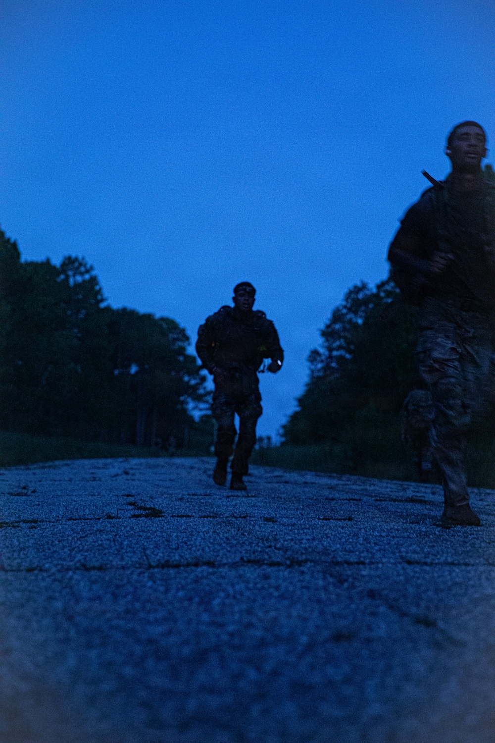 Two military personnel in camouflage uniforms and tactical gear are running along a paved road at dusk or dawn. The low-light conditions create a deep blue cast across the scene, with tree lines visible on both sides of the road. The image appears to capture a training run or tactical movement exercise.