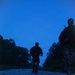 Two military personnel in camouflage uniforms and tactical gear are running along a paved road at dusk or dawn. The low-light conditions create a deep blue cast across the scene, with tree lines visible on both sides of the road. The image appears to capture a training run or tactical movement exercise.