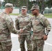 Three U.S. Army soldiers in OCP (Operational Camouflage Pattern) uniforms are engaged in a meeting or greeting in what appears to be a military installation parking area. Two of the soldiers are shaking hands while a third stands between them. Name tapes visible on two soldiers read 'CUTLER' and 'ROME', with 'U.S. ARMY' tape visible on both.