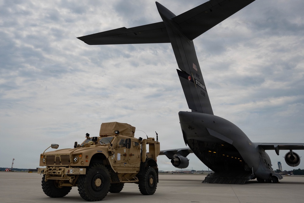 A tan/desert-colored MRAP-type military vehicle (likely an Oshkosh M-ATV) is positioned on an airfield tarmac adjacent to the open rear cargo ramp of a large gray military transport aircraft. The aircraft's tail section is prominently visible with markings reading 'STEWART' and what appears to be a unit designation. The scene depicts a military airlift or deployment operation at what appears to be a U.S. military airfield.