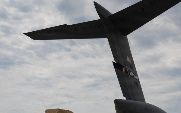 A tan/desert-colored MRAP-type military vehicle (likely an Oshkosh M-ATV) is positioned on an airfield tarmac adjacent to the open rear cargo ramp of a large gray military transport aircraft. The aircraft's tail section is prominently visible with markings reading 'STEWART' and what appears to be a unit designation. The scene depicts a military airlift or deployment operation at what appears to be a U.S. military airfield.