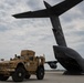 A tan/desert-colored MRAP-type military vehicle (likely an Oshkosh M-ATV) is positioned on an airfield tarmac adjacent to the open rear cargo ramp of a large gray military transport aircraft. The aircraft's tail section is prominently visible with markings reading 'STEWART' and what appears to be a unit designation. The scene depicts a military airlift or deployment operation at what appears to be a U.S. military airfield.