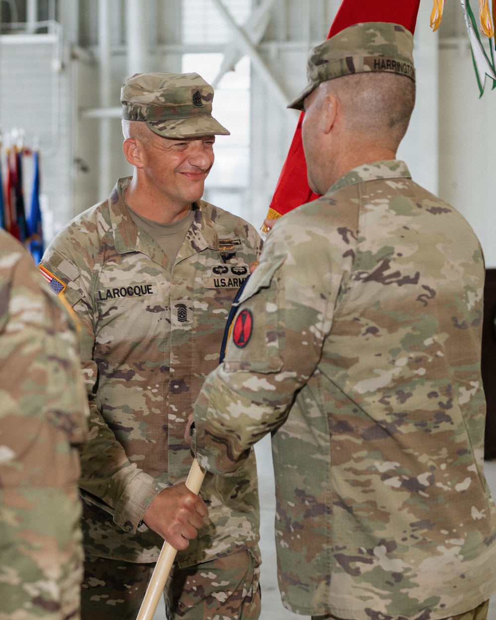 Two U.S. Army soldiers in OCP (Operational Camouflage Pattern) uniforms are engaged in what appears to be a military ceremony, likely a change of responsibility or assumption of command. The soldier facing the camera, identified by name tape as 'LAROCQUE,' holds a guidon staff and wears Command Sergeant Major rank insignia. The soldier with his back to the camera is identified by name tape as 'HARRINGTON' and both wear the 7th Army patch on their left shoulders.