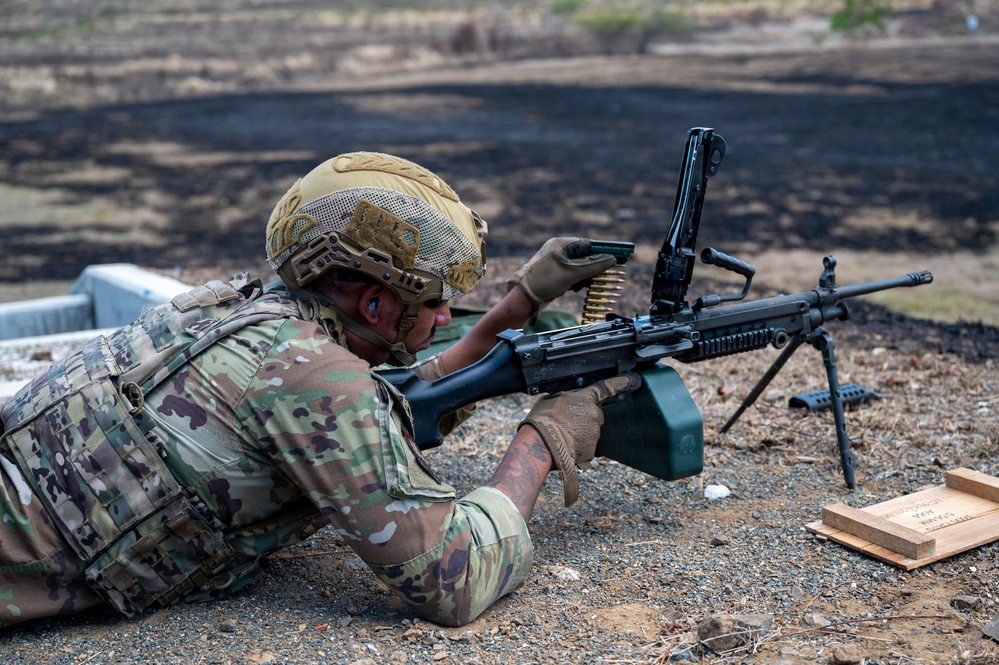 A soldier in full combat gear is in a prone firing position operating a belt-fed machine gun on a training range. The individual is wearing MultiCam/OCP camouflage uniform, a tan tactical helmet, plate carrier vest, and shooting gloves. An ammunition box is attached to the weapon and a wooden ammunition crate is visible nearby.