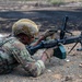 A soldier in full combat gear is in a prone firing position operating a belt-fed machine gun on a training range. The individual is wearing MultiCam/OCP camouflage uniform, a tan tactical helmet, plate carrier vest, and shooting gloves. An ammunition box is attached to the weapon and a wooden ammunition crate is visible nearby.
