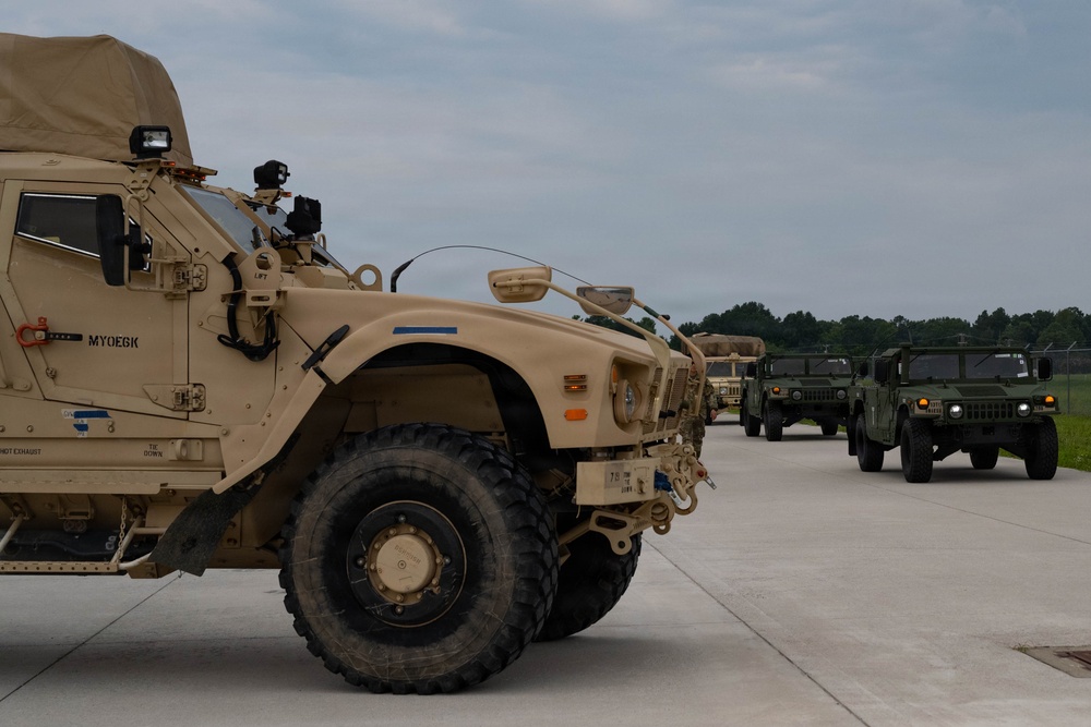 A convoy of military vehicles is staged on a concrete surface, likely a military installation or airfield. The foreground features a tan/desert-colored MRAP (Mine-Resistant Ambush Protected) vehicle, with additional vehicles including Humvees visible in the background. The vehicles appear to be lined up in preparation for movement or inspection.