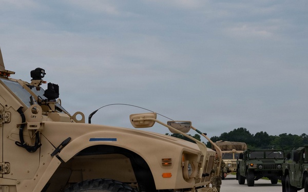 A convoy of military vehicles is staged on a concrete surface, likely a military installation or airfield. The foreground features a tan/desert-colored MRAP (Mine-Resistant Ambush Protected) vehicle, with additional vehicles including Humvees visible in the background. The vehicles appear to be lined up in preparation for movement or inspection.