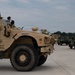 A convoy of military vehicles is staged on a concrete surface, likely a military installation or airfield. The foreground features a tan/desert-colored MRAP (Mine-Resistant Ambush Protected) vehicle, with additional vehicles including Humvees visible in the background. The vehicles appear to be lined up in preparation for movement or inspection.