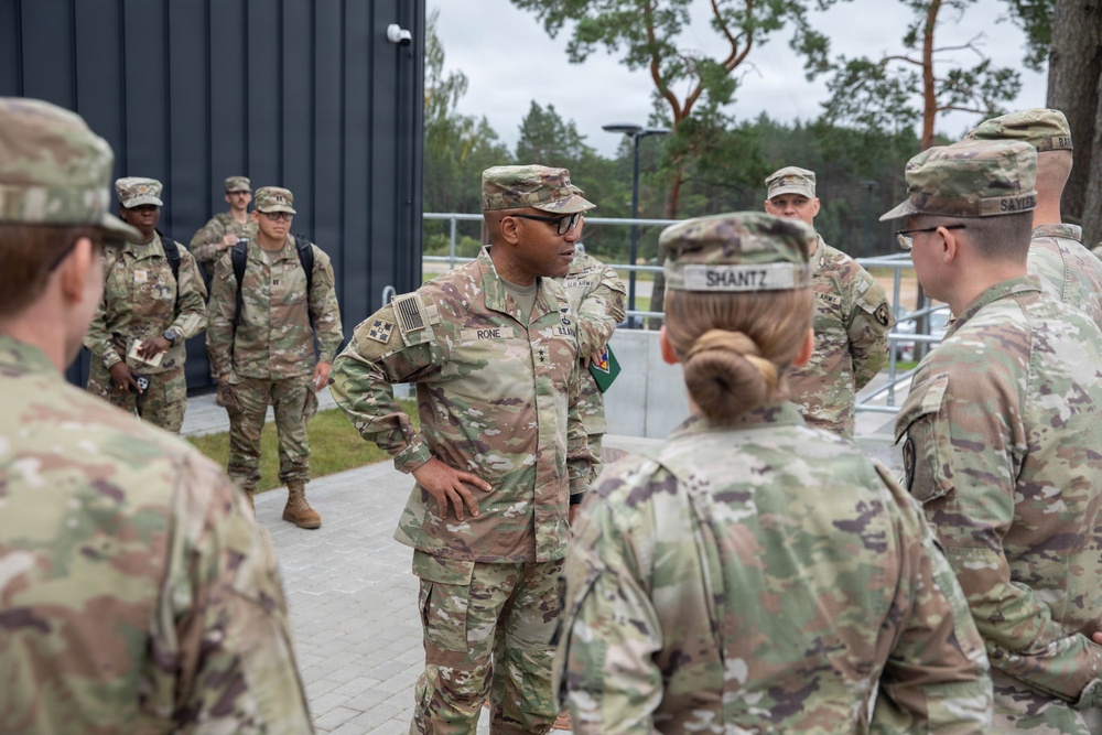 A group of U.S. Army soldiers in OCP (Operational Camouflage Pattern) uniforms are gathered outdoors near a modern building, appearing to be in a briefing or discussion. A central figure identified by nametape as 'RONE' with a general officer star visible on their chest is addressing the group. The setting appears to be a military installation, possibly in Europe based on the architecture and vegetation.