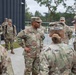 A group of U.S. Army soldiers in OCP (Operational Camouflage Pattern) uniforms are gathered outdoors near a modern building, appearing to be in a briefing or discussion. A central figure identified by nametape as 'RONE' with a general officer star visible on their chest is addressing the group. The setting appears to be a military installation, possibly in Europe based on the architecture and vegetation.