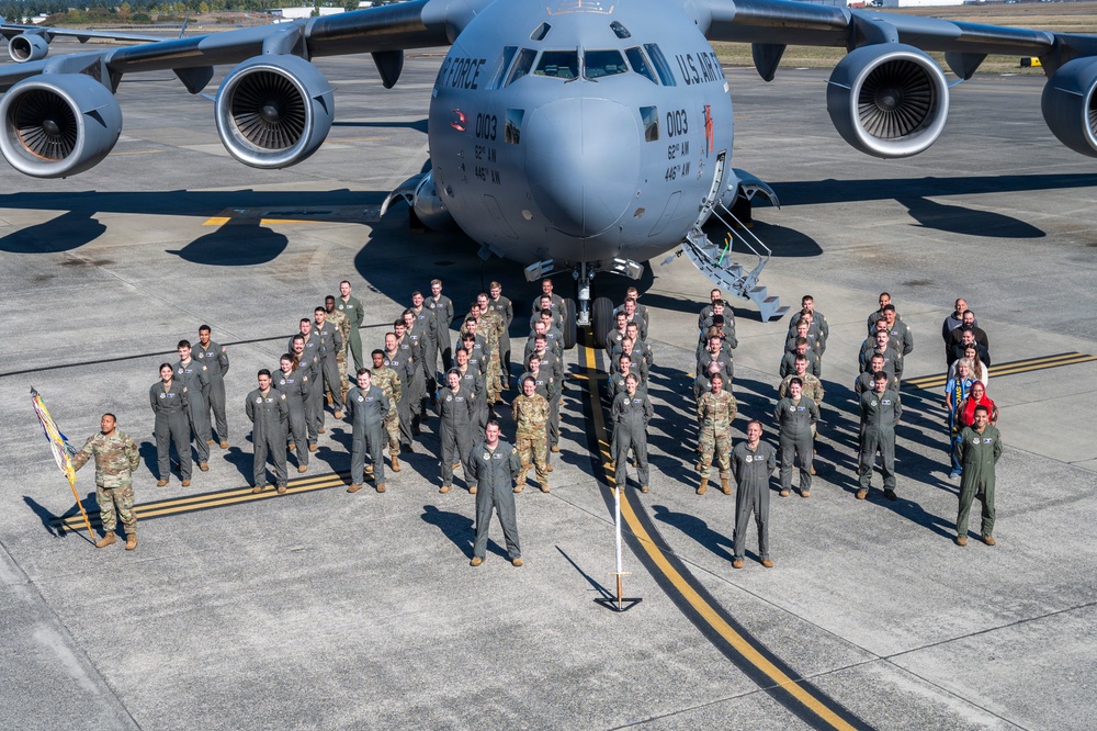 A large group of U.S. Air Force personnel pose for a formal group photograph in front of a C-17 Globemaster III military transport aircraft on a flight line tarmac. The aircraft bears the tail number 0103 and markings for the 62nd AW and 446th AW, indicating Joint Base Lewis-McChord (JBLM) in Washington State. Personnel are wearing a mix of flight suits and Army Combat Uniforms (ACU/OCP), suggesting a joint or mixed-unit gathering.