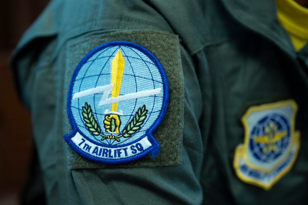 The image shows a close-up of a military flight suit sleeve bearing the patch of the 7th Airlift Squadron (7th AS), a unit of the United States Air Force. The primary patch features a globe, lightning bolt, sword, fist, and laurel wreath on a blue circular background. A second, partially visible patch on an adjacent sleeve appears to be a USAF command or wing insignia.