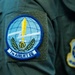 The image shows a close-up of a military flight suit sleeve bearing the patch of the 7th Airlift Squadron (7th AS), a unit of the United States Air Force. The primary patch features a globe, lightning bolt, sword, fist, and laurel wreath on a blue circular background. A second, partially visible patch on an adjacent sleeve appears to be a USAF command or wing insignia.