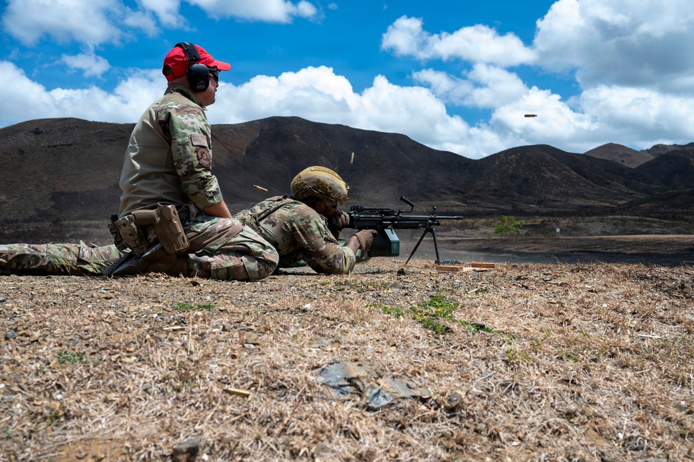 Two military personnel in MultiCam/OCP camouflage uniforms are conducting a live-fire weapons training exercise at an outdoor range. One soldier is in the prone position firing a light machine gun (appears to be an M249 SAW or M240B on a bipod), while a second individual wearing a red cap and hearing protection observes and supervises. Spent brass casings are visibly ejecting from the weapon, captured mid-air by the camera.