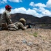 Two military personnel in MultiCam/OCP camouflage uniforms are conducting a live-fire weapons training exercise at an outdoor range. One soldier is in the prone position firing a light machine gun (appears to be an M249 SAW or M240B on a bipod), while a second individual wearing a red cap and hearing protection observes and supervises. Spent brass casings are visibly ejecting from the weapon, captured mid-air by the camera.