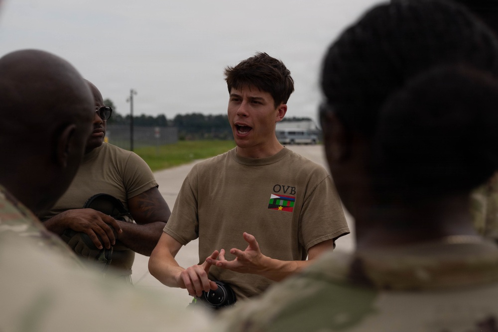 A young man wearing a tan/khaki t-shirt with 'OVB' text and a small flag graphic is speaking animatedly to a group of individuals in military uniforms. The setting appears to be an outdoor military installation or airfield, with a tarmac or road visible in the background. The speaker appears to be briefing or explaining something using hand gestures.