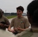 A young man wearing a tan/khaki t-shirt with 'OVB' text and a small flag graphic is speaking animatedly to a group of individuals in military uniforms. The setting appears to be an outdoor military installation or airfield, with a tarmac or road visible in the background. The speaker appears to be briefing or explaining something using hand gestures.