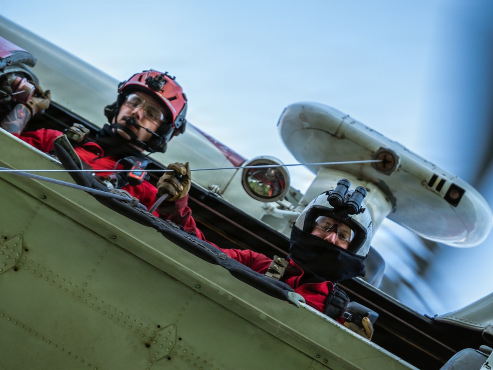 Two individuals in red flight suits are seated in an open cockpit of a vintage or classic aircraft, photographed from a low angle during flight. Both wear helmets and protective eyewear, with one wearing a red helmet and the other a white helmet equipped with what appears to be a monocular or optical device. The aircraft's fuselage, bracing wires, and a spinning propeller are visible in the background.