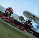 Two individuals in red flight suits are seated in an open cockpit of a vintage or classic aircraft, photographed from a low angle during flight. Both wear helmets and protective eyewear, with one wearing a red helmet and the other a white helmet equipped with what appears to be a monocular or optical device. The aircraft's fuselage, bracing wires, and a spinning propeller are visible in the background.