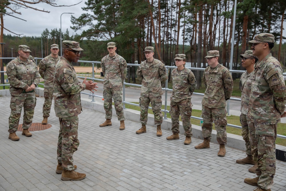 A senior U.S. Army soldier, likely a non-commissioned or commissioned officer, addresses a group of approximately nine soldiers in an outdoor briefing or informal formation. All personnel are wearing U.S. Army OCP (Operational Camouflage Pattern) uniforms with patrol caps. The setting appears to be a military installation or training area with a paved courtyard, metal railing, and forested background.