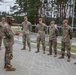 A senior U.S. Army soldier, likely a non-commissioned or commissioned officer, addresses a group of approximately nine soldiers in an outdoor briefing or informal formation. All personnel are wearing U.S. Army OCP (Operational Camouflage Pattern) uniforms with patrol caps. The setting appears to be a military installation or training area with a paved courtyard, metal railing, and forested background.