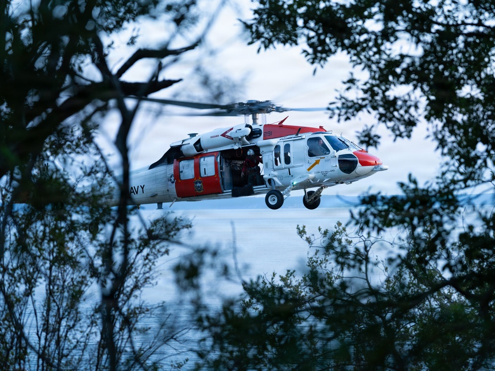 A U.S. Navy search and rescue helicopter, identifiable by its white and red color scheme and 'NAVY' markings, is captured in low-level flight framed through tree branches near a body of water. The aircraft appears to be a Sikorsky MH-60S Knighthawk or similar variant, with its side door open and a crew member visible inside. The image is taken at dusk or dawn, giving the scene a cool blue tone.